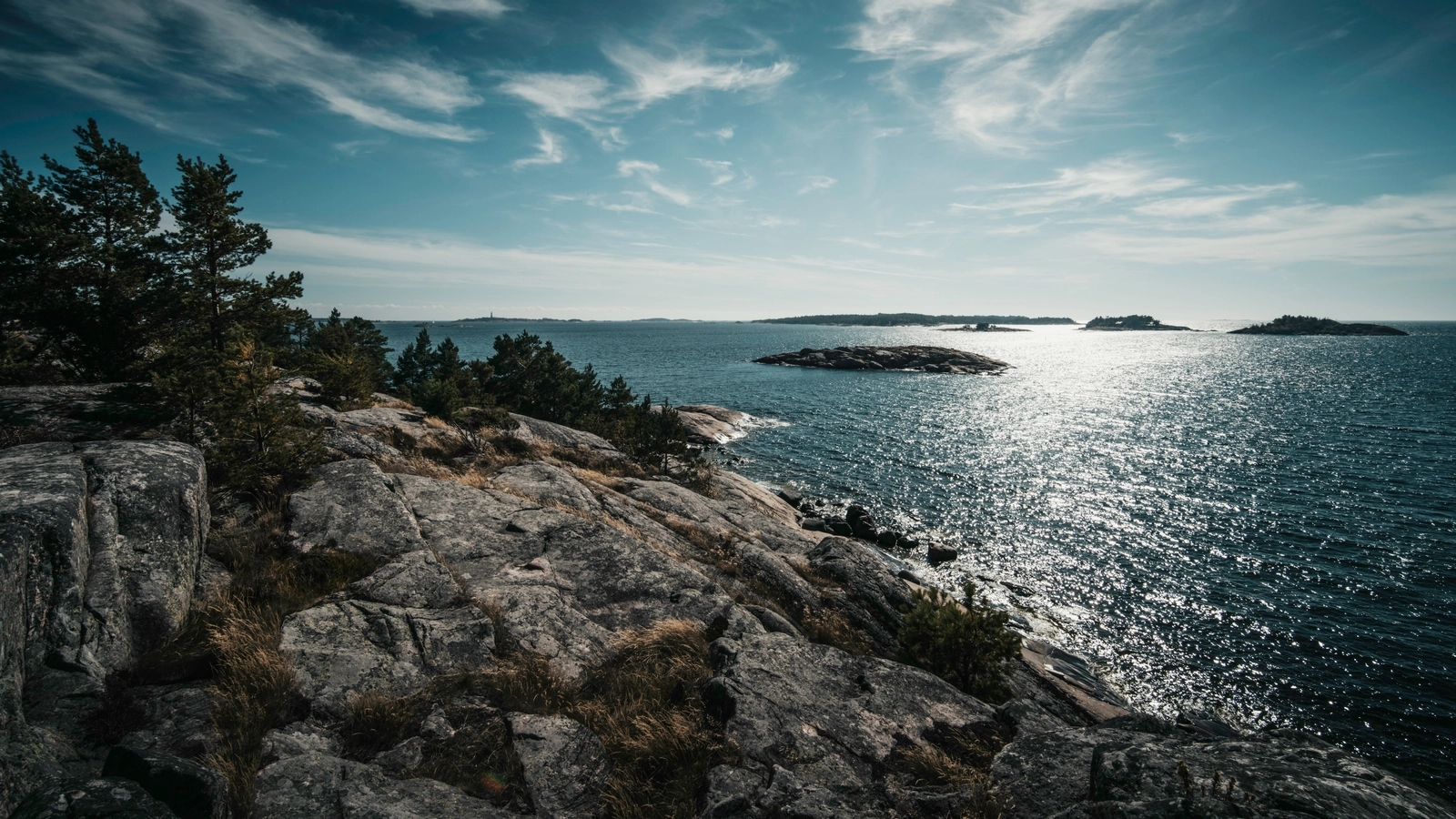 Rocky coastline with pine trees overlooking a sunlit, sparkling blue sea dotted with small islands under a partly cloudy sky.